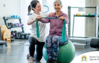 A care provider helps an older woman exercise with a resistance band and an exercise ball, representing how exercise can help with senior fall prevention.